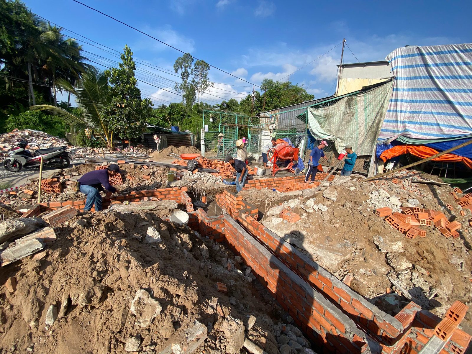 Nhiều thợ đang xây móng gạch đỏ trong các rãnh, có máy trộn bê tông và xe rùa