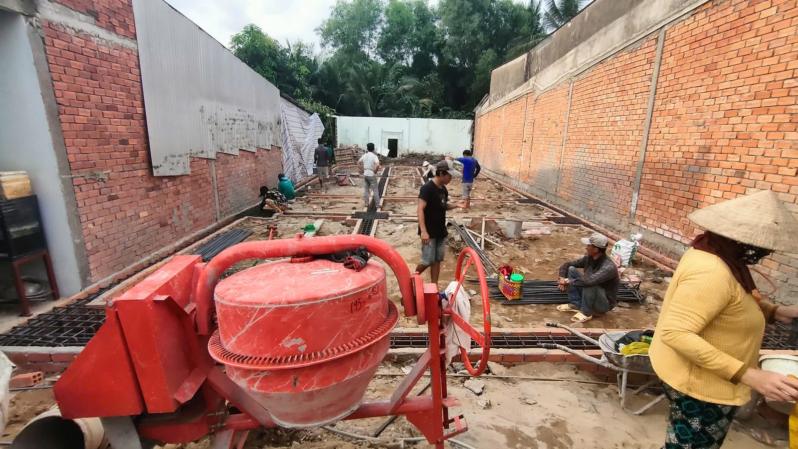 Máy trộn bê tông đỏ ở tiền cảnh, phía sau tổ thợ đang lắp hệ thép đà kiềng